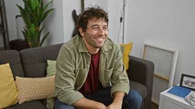 Güzel dekore edilmiş bir oturma odasında gri bir kanepede oturan rahat ceketli, gülümseyen genç bir İspanyol..