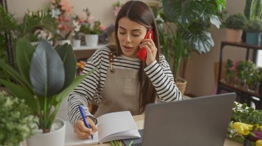 Evdeki defterde yazarken telefonda konuşan İspanyol kadın. Etrafı bitkilerle çevrili..