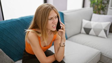 Modern oturma odasında akıllı telefondan konuşan sinirli genç bir kadın, sarı saçlı ve anlamlı yüzlü..