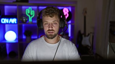 Geceleri neon ışıklı karanlık oyun odasında kulaklık takan mavi gözlü sakallı genç bir adam.