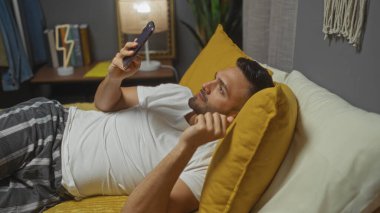 Hispanik bir adam, yatak odasında akıllı telefon kullanarak yatağında uzanıyor ve rahat bir ev ortamını gösteriyor..