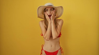 Kırmızı bikinili ve güneş şapkalı çekici genç bir kadın sarı arka planda poz veriyor, iki eli de suratında şaşırdığını ifade ediyor..