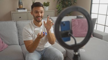 Oturma odasında oturan sakallı, elinde bir konteynır, gülümseyen ve elinde akıllı telefonuyla video kaydeden genç bir İspanyol..