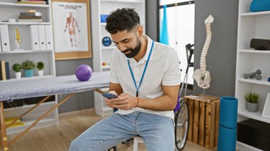 İyi donanımlı bir kliniğin rehabilitasyon odasında akıllı telefondan mesaj atan sakallı yakışıklı bir genç.