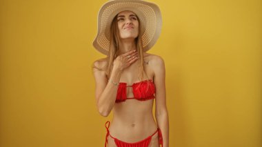 Kırmızı bikinili ve geniş kenarlı şapkalı güzel bir kadın izole edilmiş sarı arka plan duvarına karşı boğazını acı içinde tutuyor..