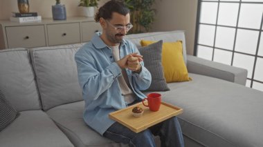 Bıyıklı genç İspanyol bir adam oturma odasında oturmuş ellerini tahta bir tepsiyle ovuşturuyor ve kucağında kırmızı bir fincan ve kek tutuyor.