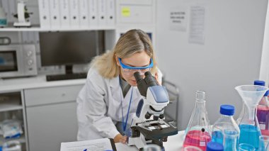 Beyaz kadın bilim adamı, kapalı bir laboratuvar ortamında mikroskopla örnekleri analiz ediyor.