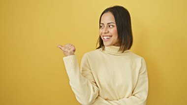 Bej kazaklı genç İspanyol kadın gülümsüyor ve sarı arka planı işaret ediyor.