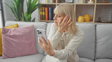 Sarışın kadın oturma odasının iç tarafındaki kanepede akıllı telefon kullanıyor.