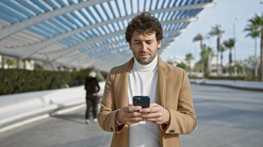 Yakışıklı İspanyol ceketli adam modern mimariyle güneşli şehir gezintisinde akıllı telefon kullanıyor.