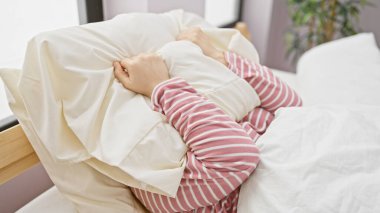 Çizgili pijamalı genç bir kadın rahat ve rahatlatıcı duygular uyandıran rahat bir yatak odasında yastığa sarılıyor..