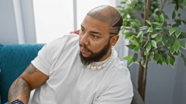 Sakallı şık bir Afrikalı Amerikalı modern bir oturma odasında düşünceli bir şekilde poz veriyor, her zamanki gibi zarif bir hava yayıyor..