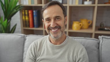 Orta yaşlı neşeli sakallı bir adam modern bir oturma odasında oturuyor ve arka planda kitap rafı ve çiçekleri var..