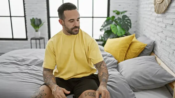 stock image A young, bearded man sits thoughtfully on a bed in a well-lit, modern room, exuding a sense of casual style and comfort.