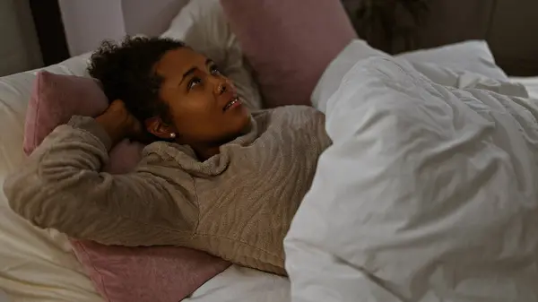 stock image A contemplative young woman with curly hair relaxes in her cozy bedroom, looking thoughtful and serene.