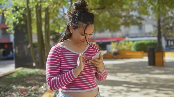 stock image A young hispanic woman in casual attire uses her smartphone outdoors in a sunny urban park.