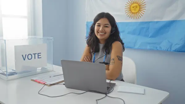 stock image Young hispanic woman inside an argentinian electoral college room, seated at a desk with a laptop, in front of a voting ballot box, with an argentine flag in the background.