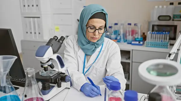 stock image A mature woman scientist in hijab meticulously taking notes in a laboratory setting.