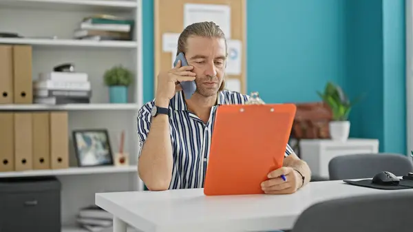 stock image A professional man multitasks with a phone and clipboard in a modern office environment.