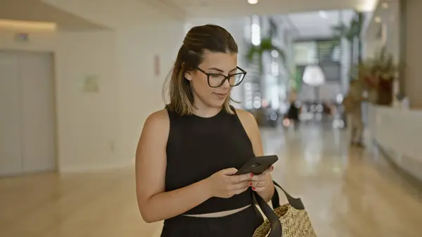 stock image Beautiful adult hispanic woman using her smartphone in a dynamic hotel hall - a girl in glasses connected, typing a serious text in digital comfort