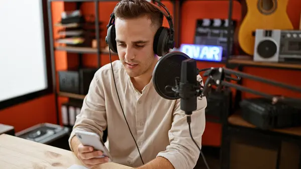 stock image Handsome young man with headphones in a music studio looking at smartphone, equipment visible