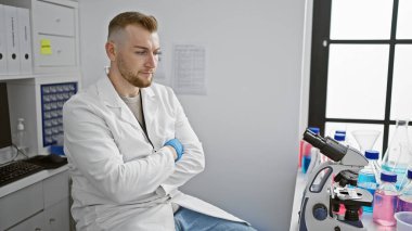 Laboratuvar önlüklü düşünceli bir adam mikroskop ve laboratuvar malzemelerinin yanında modern bir laboratuvarda oturuyor..