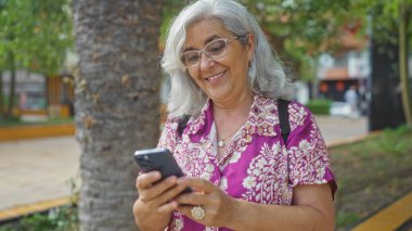Şehir parkında akıllı telefon kullanan yaşlı bir kadın gülümsüyor ve güneşli bir günün tadını çıkarıyor.