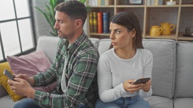 Bir adam ve bir kadın, rahat bir oturma odasındaki kanepede yan yana otururken dikkatlerini dağıtarak telefonlarını kullanıyorlar..