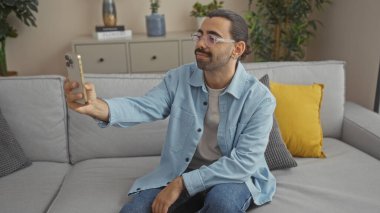 Bıyıklı genç bir adam bir apartman dairesinde selfie çekiyor, gözlüklü ve açık mavi gömlekli, arka planda sarı yastıklı ve kapalı alanda bitkiler olan bir kanepede oturuyor.