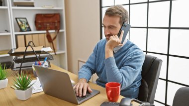 Orta yaşlı İspanyol bir adam, iyi aydınlatılmış bir ofis ortamında dizüstü bilgisayar kullanırken bir telefon görüşmesi yapıyor..