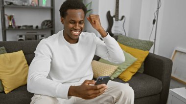 Genç Afrikalı Amerikalı adam, modern apartman dairesinde başarıyı akıllı telefonla kutluyor.