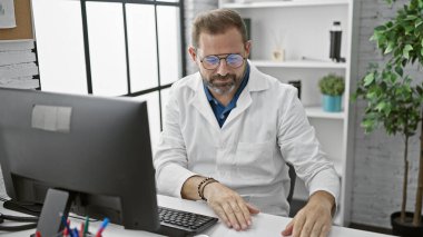 Yakışıklı, beyaz önlüklü olgun bir adam modern bir klinikteki bir bilgisayarda çalışıyor..