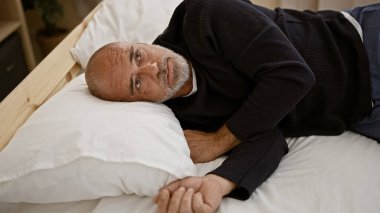 Olgun, kel, sakallı bir adam yatakta uzanıyor, rahat bir yatak odası ortamında kameraya bakıyor..