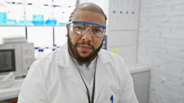 Sakal ve güvenlik gözlüğü takan kendine güvenen siyah bir adam laboratuarda beyaz önlüklü poz veriyor..