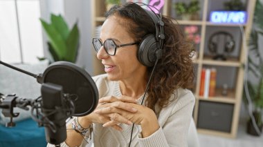 Gülümseyen, yetişkin, kulaklık takan İspanyol bir kadın modern bir radyo stüdyosunda mikrofona konuşuyor..