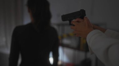 Silahlı bir kişi, loş bir mekanda bir kadının sırtına silah doğrultuyor. Gergin bir yüzleşme ima ediyor..