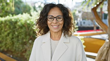 Güneşli bir parkta duran, gözlüklü, beyaz ceketli, gülümseyen olgun İspanyol kadın.