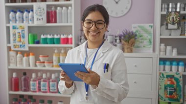Eczanede beyaz laboratuvar önlüğü ve gözlük takan gülümseyen genç bir kadın ilaç ve ürünlerle dolu rafların önünde duran bir tablet tutuyor..