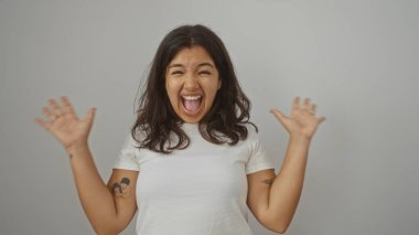 Genç, esmer, İspanyol bir kadın heyecanla gülümsüyor ve ellerini beyaz arka plan duvarına doğru kaldırıyor..