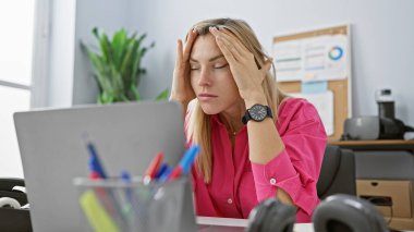 Ofisteki stresli genç kadın dizüstü bilgisayarla çalışırken baş ağrısı çekiyor, iş yorgunluğunu gösteriyor..