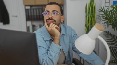 Gözlüklü ve bıyıklı İspanyol bir adam ofiste oturmuş, elleri çenesinde, etrafı iç mekan bitkileriyle ve ofis malzemeleriyle çevrili olduğunu düşünüyor.