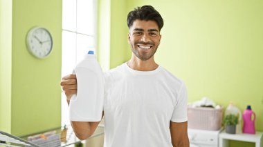 Parlak bir çamaşır odasında çamaşır deterjanı tutan yakışıklı genç adam kameraya gülümsüyor..