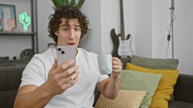 Genç adam rahat bir oturma odasında akıllı telefonunu karıştırırken kahveden zevk alıyor..
