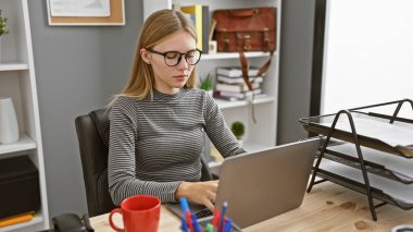 Modern bir ofis masasında dizüstü bilgisayarda çalışan, kahve fincanı ve kırtasiyesiyle odaklanmış sarışın bir kadın..