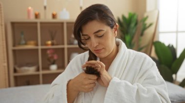 Kaplıca odasında saçları inceleyen genç bir kadın, arkasında iç mekan bitkileri ve raflarla bir sağlık merkezinde son buluyor.