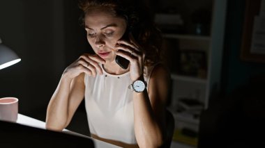 Kolsuz tişörtlü, odaklanmış, beyaz bir kadın gece yarısı ofis masasında bir telefon görüşmesi yapmakla meşgul..