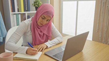 Bilgisayarının yanına parlak, modern bir ofis ortamında tesettüre yazı yazan odaklanmış bir kadın..