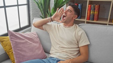 Parlak bir oturma odasında gri bir kanepede otururken İspanyol adam mutlu bir şekilde bağırıyor.