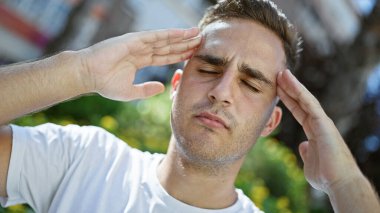 Beyaz tişörtlü genç bir İspanyol şaşı gözlerini güneşten korur ve yeşilliklerle kaplı bir parkta..