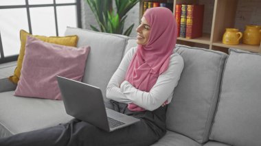 Pembe tesettürlü, gülümseyen genç bir kadın kollarını çapraz bir şekilde evdeki gri koltukta, dizüstü bilgisayarının yanında oturuyor..